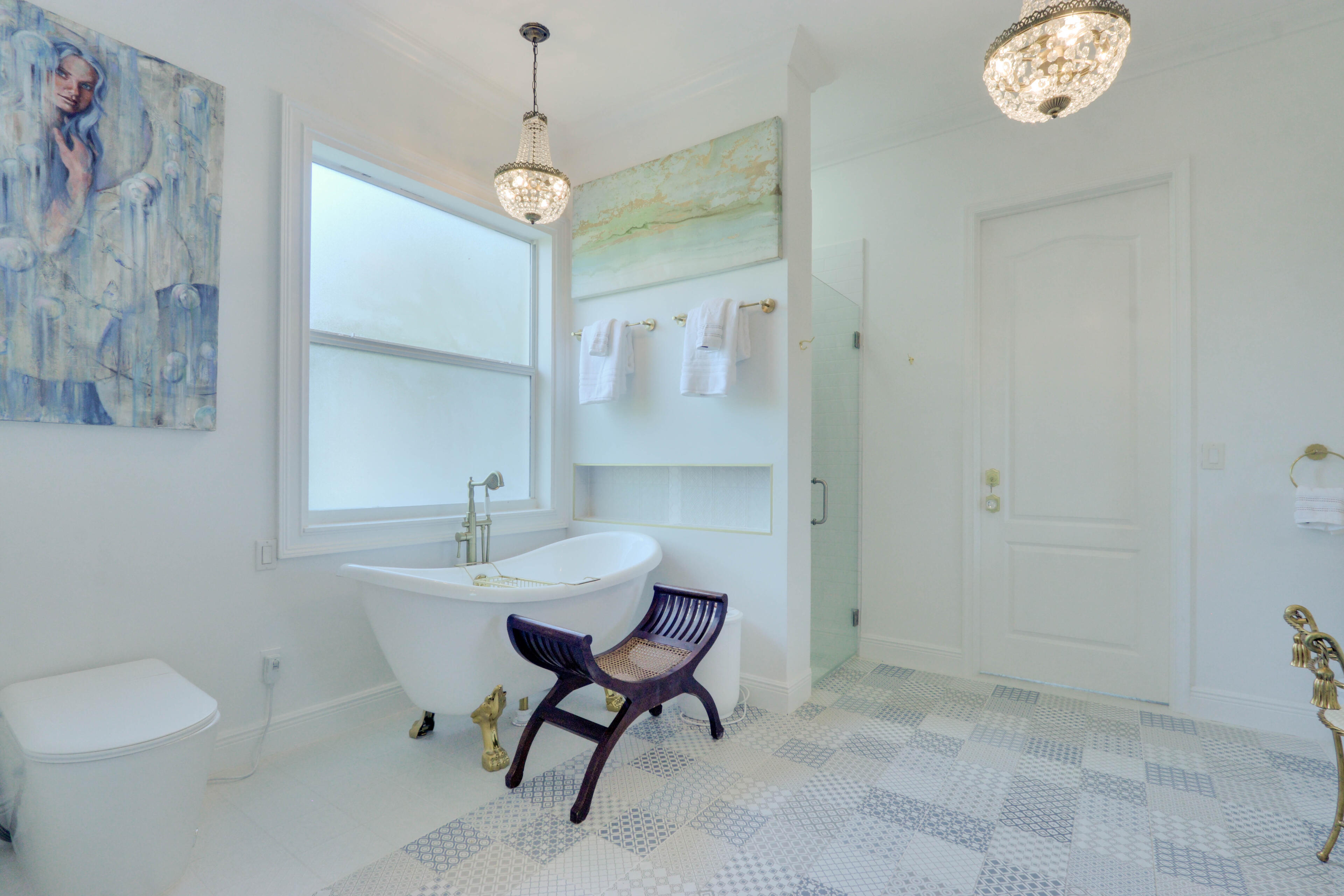 The Clawfoot Spa Bath. An oasis of classical indulgence, pairing an iconic gold-hoofed soaking tub with intricate mosaic tiling and brilliant pendant crystal lighting. Opulent white master bathroom featuring a vintage clawfoot soaking tub with gold feet, geometric mosaic tile flooring, a hanging crystal chandelier, and fine art.