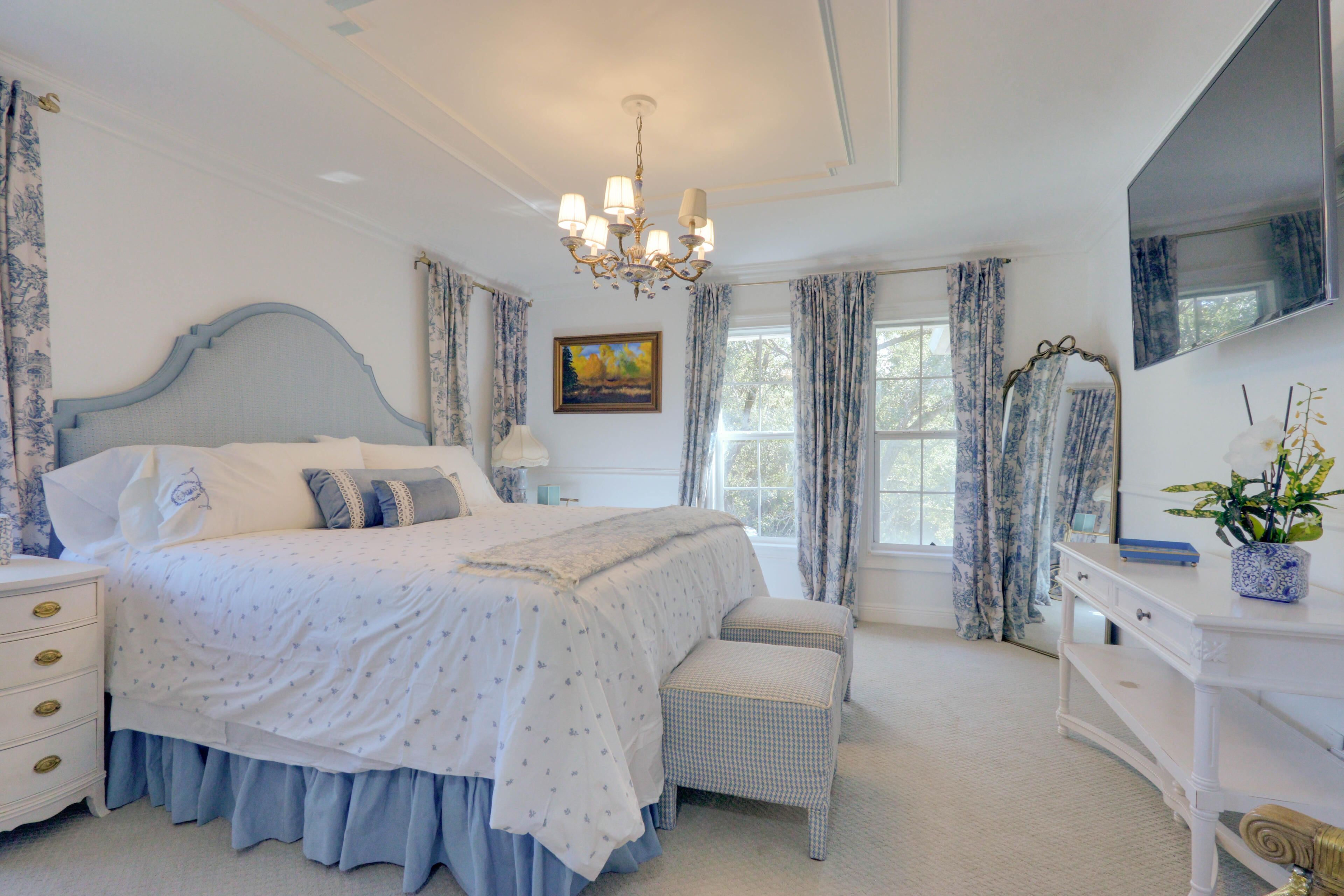 The Blue Toile Chamber. Pure Regency-core elegance draped in classical French patterns—providing an impeccably styled, romantic backdrop for period-inspired editorials. Romantic French-style bedroom with a blue upholstered headboard, matching blue toile de Jouy curtains and bedding, an ornate gold full-length mirror, and a crystal chandelier.