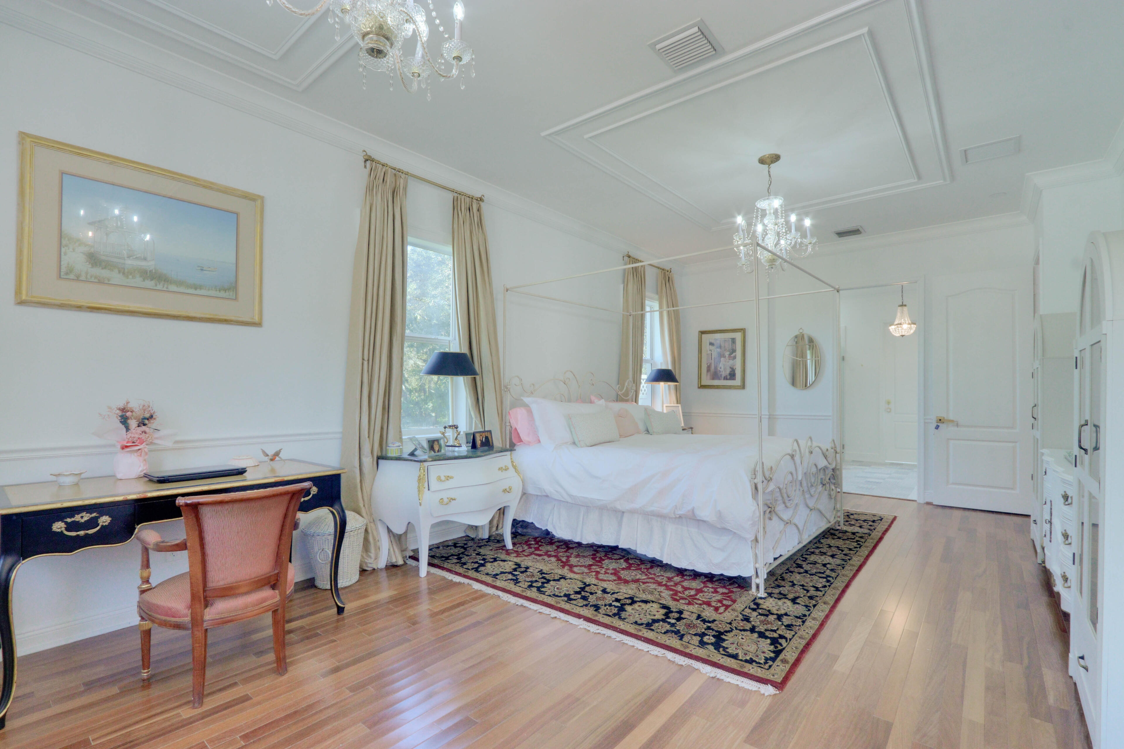 The Grand Canopy Suite. An expansive, sun-lit sanctuary anchoring the upper level, complete with a majestic iron frame and timeless European accents. Bright luxury master bedroom featuring a white iron four-poster canopy bed, dark ornate oriental rug over hardwood floors, and a crystal chandelier.