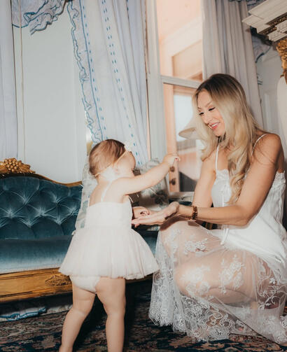 LIVING IN MOTION // Capturing candid, unscripted familial joy against rich, high-contrast jewel tones. A mother dressed in a white silk slip shares a tender moment holding hands with her standing baby girl on a vintage rug in front of a blue velvet and gold sofa.