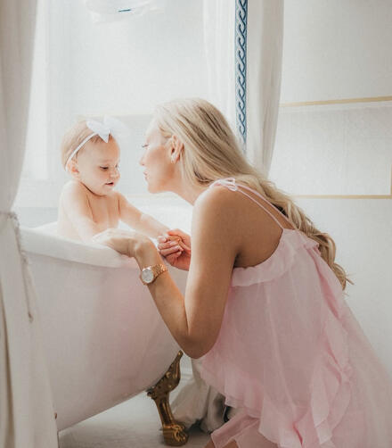 THE SUITE ATELIER // Ethereal morning light reflecting across premium porcelain bath fixtures. A mother in a soft pink ruffled dress kneels next to a vintage white clawfoot bathtub with gold legs, holding hands with her baby girl during a bright, airy family portrait session at Sarti Luxe Estates.