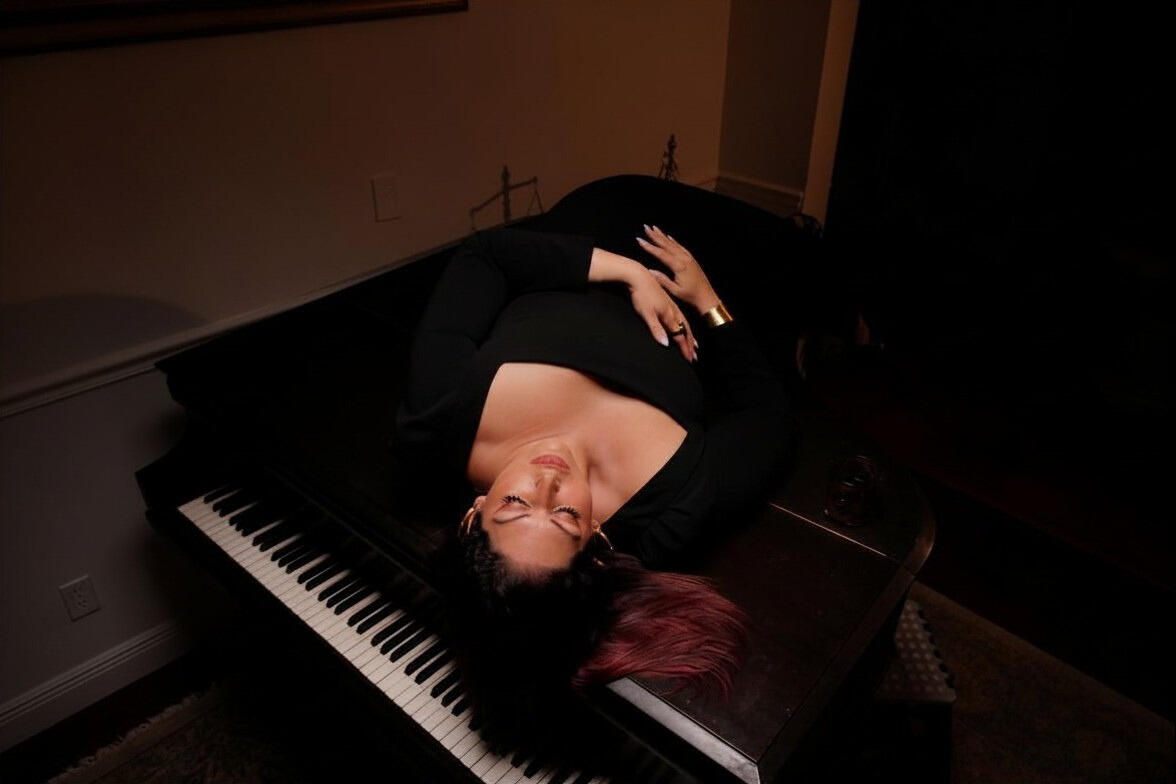 Sequin Cape on Piano Luxury editorial portrait of a woman in a sequined black cape sitting on a classic black grand piano under dramatic warm lighting.