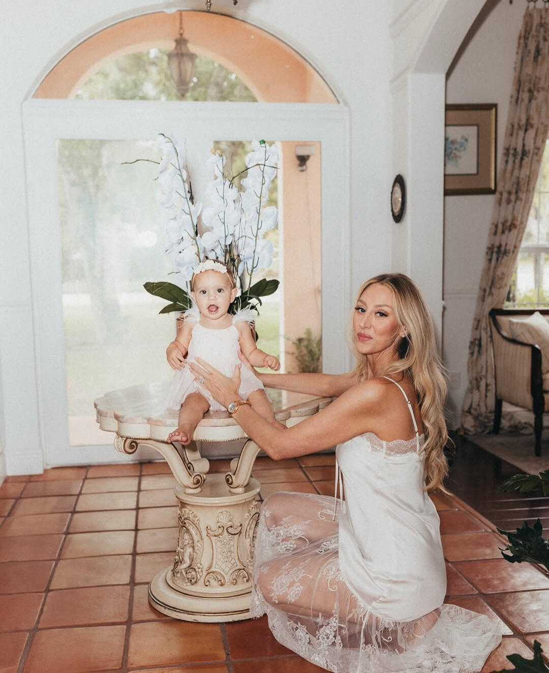 REFINED CORNERS // Every structural detail curated for flawless framing. A luxury portrait of a mother kneeling next to a marble pedestal table with fresh white orchids inside a private estate shoot location.